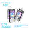 Werbebild für Airmez Fox Vape-Geräte mit zwei Vape-Einheiten im Vordergrund, einem Cartoon-Fuchs in einer blauen Mütze und einem Hemd mit farbenfrohen Spritzern im Hintergrund und Text, das 2-in-1-Aroma-Optionen hervorhebt, 8000 Puffs und Blue Razz Lemonade und Blue Razz.